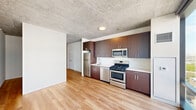 Located at 51 East 14th Street in Chicago's vibrant South Loop neighborhood, this modern studio offers an efficient and thoughtfully designed single-floor layout. The space is defined by its striking exposed concrete ceiling, which lends an industrial-chic character throughout, complemented by warm, light-toned hardwood flooring that runs seamlessly from room to room.

The open kitchen is a standout feature, outfitted with rich dark wood cabinetry extending to the ceiling for ample storage, sleek white countertops, and a full suite of stainless steel appliances including a gas range, microwave, dishwasher, and refrigerator. Floor-to-ceiling windows flood the kitchen and living area with abundant natural light while framing impressive views of the surrounding Chicago cityscape.

A hallway connects the main living area to the private spaces, including a clean, minimalist bathroom featuring a wide vanity with integrated sink, light wood cabinetry, and tiled walls. A closet and in-unit storage add to the unit's practicality. The open living area, with its crisp white walls and concrete ceiling, provides a versatile blank canvas. This studio combines urban industrial design with modern finishes, making it an ideal urban retreat in the heart of Chicago.