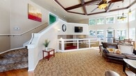 Located at 2820 Tuscany Ln SW in Tumwater, Washington, this single-floor community space offers an impressive blend of architectural character and functional design. The grand entryway sets the tone immediately, featuring elegant tile flooring with a diamond accent pattern, soaring ceilings with an arched transom window above the front door, and a decorative chandelier that adds warmth to the welcome area. A solid wood interior door and glass-paneled entry doors contribute to the refined aesthetic.

The main living area is defined by a striking coffered ceiling with rich dark wood beams and a ceiling fan, complemented by plush carpeting with a floral pattern. An interior railing system creates a split-level visual effect, adding architectural depth to the open layout. The focal point of the lounge area is a full-height brick fireplace flanked by large windows that invite abundant natural light and views of the surrounding greenery.

The dining area features floor-to-ceiling windows on multiple walls, bathing the space in daylight and offering pleasant outdoor views of a natural rock landscape. The kitchen is equipped with rich dark wood cabinetry, tile countertops, open shelving, and ample workspace. Throughout the space, warm neutral tones, recessed lighting, and thoughtful design details create an inviting and well-appointed environment.