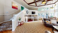 Located at 2820 Tuscany Ln SW in Tumwater, Washington, this single-floor community space offers an impressive blend of architectural character and functional design. The grand entryway sets the tone immediately, featuring elegant tile flooring with a diamond accent pattern, soaring ceilings with an arched transom window above the front door, and a decorative chandelier that adds warmth to the welcome area. A solid wood interior door and glass-paneled entry doors contribute to the refined aesthetic.

The main living area is defined by a striking coffered ceiling with rich dark wood beams and a ceiling fan, complemented by plush carpeting with a floral pattern. An interior railing system creates a split-level visual effect, adding architectural depth to the open layout. The focal point of the lounge area is a full-height brick fireplace flanked by large windows that invite abundant natural light and views of the surrounding greenery.

The dining area features floor-to-ceiling windows on multiple walls, bathing the space in daylight and offering pleasant outdoor views of a natural rock landscape. The kitchen is equipped with rich dark wood cabinetry, tile countertops, open shelving, and ample workspace. Throughout the space, warm neutral tones, recessed lighting, and thoughtful design details create an inviting and well-appointed environment.