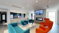 This single-floor community room is a well-appointed shared space designed to foster social interaction and comfort. The open layout seamlessly connects a lounge area with a functional kitchenette, creating a versatile environment for gatherings and everyday use.

The lounge area is anchored by a striking combination of teal upholstered sofas and armchairs paired with bold orange accent chairs, lending the space an energetic, modern aesthetic. A low white coffee table serves as a central gathering point. A wall-mounted flat-screen television is positioned above a sleek, horizontal gas fireplace framed in light mosaic tile, providing both entertainment and a warm focal point for the room.

The kitchenette runs along the back wall and features dark cabinetry, pendant lighting, a countertop with bar-height seating, and modern appliances, making it well-suited for casual dining or entertaining. Recessed lighting throughout the white ceiling ensures the space is evenly and brightly lit. Large windows allow natural light to fill the room, enhancing the clean, contemporary atmosphere. The light tile flooring ties the space together with a polished, cohesive finish.