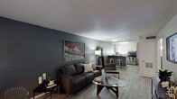 This single-floor unit showcases a distinctive industrial-inspired aesthetic, anchored by painted concrete brick accent walls that run through the living area and bedrooms. Wood-look plank flooring extends throughout the entire space, providing a cohesive and low-maintenance foundation. The open-concept living area is a standout feature, offering a generous layout that flows seamlessly into the kitchen. A dedicated workspace area with a desk sits near a bright window fitted with vertical blinds, allowing natural light to filter in. The kitchen is equipped with stainless steel appliances, a gas range, a tile backsplash, white cabinetry, and a breakfast bar with seating — ideal for casual dining. The unit includes multiple bedrooms, each carrying the same brick wall character and wood-look flooring. The bedrooms are well-proportioned, each featuring windows with vertical blinds and closets with built-in shelving and hanging rods. One bedroom features a wall-mounted flat-screen TV as a built-in detail. The textured ceilings are consistent throughout. Overall, the space presents a modern, urban feel with durable finishes, making it a functional and stylish living environment.