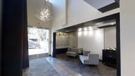 This single-floor lobby space makes a striking first impression through its dramatic double-height ceilings, which amplify the sense of openness and grandeur. A standout crystal branch chandelier serves as the focal centerpiece, casting warm light across the space and adding an artistic touch to the otherwise clean, contemporary design.

The flooring features large-format stone tiles with an intricate decorative pattern, anchoring the space with texture and visual interest. Floor-to-ceiling glass at the entrance floods the lobby with natural light, offering views of the surrounding streetscape and creating a seamless connection between the interior and exterior.

A built-in mailroom alcove sits recessed within a dark-framed niche, illuminated by track lighting and flanked by decorative bottle displays, lending a sophisticated and functional element to the space. Adjacent to the mailroom, a seating area features two modern grey upholstered chairs arranged around a small plant, providing a comfortable waiting area for residents and guests.

One wall showcases an elegant white botanical mural, adding an artistic and refined ambiance. The overall design balances modern minimalism with warm, curated details, resulting in a polished and inviting communal entrance.
