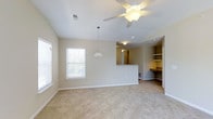 This single-story space offers an open and functional layout designed for comfortable everyday living. The main living area is finished with neutral-toned carpet and painted in a warm, light beige that creates a bright and welcoming atmosphere. Vaulted ceilings add a sense of spaciousness overhead, complemented by a ceiling fan with an integrated light fixture that provides both airflow and illumination.

A decorative pendant light hangs at the transition point between the living area and the kitchen, which is accessible via a half-wall pass-through counter. This design element maintains an open feel while defining each functional zone. The kitchen area is visible through the pass-through and features overhead lighting.

Along one wall, built-in open shelving and a built-in desk or counter surface provide practical storage and workspace options, adding a thoughtful organizational feature to the layout. Windows fitted with horizontal blinds allow natural light to filter into the living space while offering privacy control.

The overall ambiance is clean, neutral, and move-in ready, with a straightforward design that offers flexibility for a variety of furnishing arrangements and personal styles.