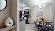This single-floor residence offers a practical and cohesive layout across several well-defined living spaces. The galley-style kitchen is efficiently organized with gray cabinetry lining both sides, dark granite-style countertops, and black appliances including a refrigerator, electric range, and over-the-range microwave. Light wood-look flooring runs throughout the kitchen, adding brightness to the space. A peninsula with a dark countertop provides a natural transition into the adjacent dining area, which features light flooring, a round wood dining table, and a window dressed with soft blue curtains that allow natural light to filter in. A chandelier overhead adds a subtle decorative touch. The living room is finished with dark wood-look flooring, neutral gray walls, and a textured ceiling, creating a relaxed and inviting atmosphere. The bedroom continues the dark flooring theme and connects directly to the hallway, offering easy access to additional rooms. The bathroom includes a full tub-shower combination with classic white subway tile surround and a curtain rod. Throughout the unit, textured walls and ceilings, neutral tones, and consistent flooring choices contribute to a cohesive, understated aesthetic.