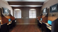 This single-floor business center is thoughtfully designed to provide a professional and functional workspace. The room features rich, dark hardwood flooring that runs throughout, complemented by warm earth-toned walls with a textured finish and crisp crown molding that adds a refined architectural detail to the space.

Natural light filters in through pairs of arched-top windows dressed with horizontal blinds, lending the room an elegant character while allowing for adjustable light control. Recessed lighting overhead ensures the space remains well-lit regardless of the time of day.

The workstation area is the focal point of the room, with several semi-private computer stations separated by tall wooden privacy dividers. Each station is equipped with a desktop computer, keyboard, and mouse, set on individual desks with ergonomic seating. Task lighting is mounted at each station for added convenience.

A built-in wooden bookshelf unit lines one wall, stocked with books and decorative accents, adding warmth and a library-like quality to the environment. Framed artwork is displayed throughout the room, enhancing the professional yet inviting ambiance. The space opens to an adjacent area, suggesting easy accessibility within a larger facility. Overall, this business center balances functionality with a polished, classic aesthetic.