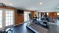 This dedicated fitness center is thoughtfully designed to support a full range of workout routines. The space is finished with warm terra-cotta-toned walls, white crown molding, and recessed lighting set against a light ceiling, creating an energizing yet polished atmosphere. Dark large-format tile flooring runs throughout, providing a durable and easy-to-maintain surface suited for active use.

The cardio zone is well-appointed, featuring Matrix treadmills and a Life Fitness elliptical trainer, complemented by a wall-mounted flat-screen television for entertainment during workouts. French doors on multiple walls allow ample natural light to filter in and offer views of the surrounding exterior. Additional windows further brighten the space and enhance ventilation.

The strength training area is equally comprehensive, with a multi-station cable and weight machine serving as the centerpiece, alongside a dedicated free weights rack holding a full set of dumbbells and an adjustable bench for varied exercises. Full-length mirrors line the walls, visually expanding the space while allowing users to monitor their form. A water fountain is also conveniently located within the room. The overall layout is open and functional, offering sufficient floor space for stretching and floor-based exercises, making this a complete and versatile fitness facility.