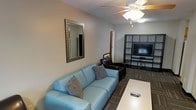 This single-floor space offers a comfortable and functional living area designed with a clean, modern aesthetic. The main living room is furnished with a light blue leather sofa accented by decorative throw pillows, complemented by a dark leather armchair, creating a welcoming seating arrangement. A rectangular white coffee table anchors the space, providing both style and practicality.

A ceiling fan with an integrated light fixture is centrally positioned overhead, ensuring air circulation and ambient lighting throughout the room. Along one wall, a large dark-framed open shelving unit houses a flat-screen television alongside organized storage compartments, serving as the focal point of the entertainment area. A framed rectangular mirror with a sleek metallic border adorns the adjacent wall, adding depth and visual interest to the room.

The flooring features a patterned carpet with earthy tones, lending warmth to the space, while the neutral-toned walls provide a versatile backdrop for the furnishings. A doorway visible toward the back of the room suggests additional spaces beyond the main living area. Overall, the space presents a well-organized, inviting environment suited for everyday living and relaxation.