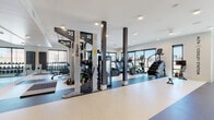 The Alta Cooley Station fitness center is a thoughtfully designed, single-floor facility that offers a comprehensive workout environment for residents. The space is organized into distinct functional zones, each tailored to a specific type of training. The open-concept layout is anchored by dark structural columns that define the space without obstructing sightlines, contributing to an airy and expansive feel throughout.

The cardio zone is positioned along a wall of large, floor-to-ceiling windows that flood the area with natural light and provide open outdoor views. This section is equipped with multiple treadmills, elliptical machines, and a stair climber, all featuring digital displays for performance tracking. Adjacent to this area, a dedicated row of upright exercise bikes offers additional low-impact cardio options.

The strength training section features an Octagon multi-functional cable and resistance system, a full rack of dumbbells displayed on tiered shelving, and a selection of selectorized weight machines for targeted muscle group training. A floor-to-ceiling mirror spans one wall, enhancing the sense of space and providing visual feedback during workouts.

The flooring transitions between speckled rubberized surfaces in light and dark tones, delineating each training zone clearly. Recessed ceiling lighting ensures consistent illumination throughout. The overall aesthetic is modern and clean, with white walls and a neutral palette that gives the facility a polished, professional character.