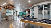 This expansive single-floor amenity space is thoughtfully designed to accommodate a variety of social and recreational activities. The open-concept layout features polished concrete floors throughout, lending an industrial-modern aesthetic that ties the zones together cohesively.

The central lounge area serves as the heart of the space, anchored by a sleek fireplace with a wood mantel and a wall-mounted flat-screen TV above it. Flanking textured accent walls and a coffered ceiling with dark-painted recessed panels add dimension and visual interest. Comfortable seating, including neutral sofas and bold blue accent chairs, creates an inviting atmosphere for relaxation or socializing.

Adjacent to the lounge, a billiards table occupies a generously sized game zone beneath striking suspended wood-panel ceiling accents. The kitchenette area is well-appointed with stainless steel appliances, white cabinetry, stone countertops, pendant lighting, and a center island with bar seating — ideal for casual entertaining. A coffee station and full-size refrigerator enhance the functionality of this area.

A café-style dining zone features high-top round tables with bistro chairs and a wall-mounted TV, creating a comfortable space for informal gatherings. Floor-to-ceiling glass partition walls connect this area to an adjacent fitness room, maintaining visual openness while providing acoustic separation. Recessed lighting throughout ensures the entire space is well-lit and welcoming.