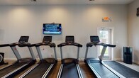 Located at 5695 Cherry Bottom Rd in Columbus, Ohio, this fitness center offers a comprehensive workout facility designed to support a wide range of exercise routines. The space is laid out on a single floor with a clean, modern aesthetic, featuring neutral-toned walls, recessed lighting, and durable carpeted flooring throughout.

The cardio section is a central highlight, housing a row of treadmills and elliptical trainers, each equipped with individual digital consoles. Wall-mounted flat-screen televisions are positioned throughout the room, keeping exercisers entertained during their sessions. A dedicated strength training area includes an adjustable workout bench, a rack of dumbbells spanning multiple weight increments, and a cable machine, providing ample options for resistance training.

A functional training corner rounds out the space, stocked with stability balls, medicine balls, a sandbag, and foam ottomans for dynamic and flexibility-focused workouts. Large motivational fitness photographs are displayed on the walls, adding energy to the environment. Natural light filters in through windows and a glass-paneled door that offers views of the surrounding outdoor area. The facility also features clearly posted gym rules and multiple exit access points, ensuring a safe and organized environment for all users.