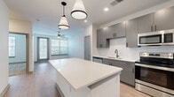 This thoughtfully designed one-bedroom, one-bathroom residence offers a cohesive blend of modern aesthetics and functional living across a single floor. The open-concept layout seamlessly connects the kitchen and living room, creating an inviting and efficient use of space. The kitchen is a standout feature, showcasing sleek gray cabinetry with matte black hardware, a striking white hexagonal tile backsplash, quartz countertops, and a large central island with pendant lighting overhead. Stainless steel appliances — including a refrigerator, range, microwave, and dishwasher — complete the contemporary look. Light wood-style flooring flows throughout the main living areas, complementing the clean, neutral color palette. The living room benefits from natural light through large windows with blinds and is accented by a ceiling fan. The bedroom offers plush carpeting, a ceiling fan, and a generously sized window that brings in natural light. A dedicated walk-in closet with wire shelving provides ample storage. The bathroom features light wood-style flooring, a dual-sink vanity with gray cabinetry, and a tiled tub-shower combination. Throughout, the space maintains a bright, modern ambiance with clean lines and quality finishes.