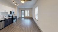 This studio is designed to maximize functionality within a thoughtfully organized single-level floor plan. The open-concept living and kitchen area is anchored by durable wood-style plank flooring that runs throughout the unit, creating visual continuity and a warm, contemporary aesthetic. Ceiling fans are present in both the living area and along the hallway corridor, contributing to comfort and air circulation.

The kitchen is a standout feature, showcasing a two-tone design with crisp white upper cabinetry and dark lower cabinets paired with granite-style countertops and a textured tile backsplash. A full suite of stainless steel appliances includes a side-by-side refrigerator with an ice and water dispenser, an electric range, over-the-range microwave, and a dishwasher. Track lighting above the kitchen provides focused illumination.

The living area benefits from natural light through windows fitted with horizontal blinds, and a set of glass doors opens to an outdoor access point. The bathroom features wood-style flooring consistent with the rest of the unit, a granite-topped dark vanity, and a full tub-shower combination with clean white tile surround and brushed nickel fixtures.