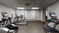 Located at 1380 E Hyde Park Blvd in Chicago's Hyde Park neighborhood, this dedicated fitness center offers a comprehensive workout facility across a single level. The space features a clean, functional design with neutral gray walls, a drop ceiling with recessed panel lighting, and durable rubber-textured flooring suited for high-traffic exercise use.

The facility is thoughtfully organized to accommodate a wide range of workout routines. Cardio equipment includes multiple True-brand treadmills, elliptical trainers, and a stationary cycle, all positioned for comfortable use. A multi-station cable and resistance machine provides options for full-body strength training, while a dedicated free weights section features a rack of dumbbells and a flat workout bench. Additional benches and a leg press machine round out the strength training area.

A large framed graphic mural of an athletic figure in motion serves as a focal point on one wall, adding a motivating visual element to the environment. The space is well-lit throughout with bright overhead fluorescent panels, ensuring clear visibility across all workout zones. Additional amenities include a sanitizing wipe dispenser, a waste bin, and safety equipment mounted on the wall. Access to adjacent rooms, likely locker or utility areas, is provided through interior doors at the rear of the facility.
