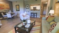This single-story space showcases an open-concept layout that connects the living area and kitchen, creating a cohesive and functional flow. The living area is anchored by a dark-framed glass coffee table atop a striped area rug in warm earth tones, complemented by light-upholstered seating and tasteful wall art that adds color and character to the neutral walls. Hardwood-style flooring extends throughout, lending warmth and visual continuity across the space. A dedicated dining nook is visible to the side, providing a defined yet integrated area for meals. The kitchen features white cabinetry, stainless steel appliances including a microwave, and a mosaic tile backsplash that serves as a decorative focal point. A raised breakfast bar with dark bar stools bridges the kitchen and living area, making it ideal for casual dining or entertaining. The overall ambiance is bright, welcoming, and thoughtfully styled, with a clean, modern aesthetic achieved through a consistent neutral color palette and carefully chosen furnishings and décor.