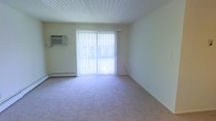 This single-floor residential space offers a clean, functional layout with a straightforward and practical design. The main living area is finished with wall-to-wall carpeting in a neutral tone, creating a warm and comfortable feel underfoot. A large sliding glass door serves as the focal point of the room, allowing an abundance of natural light to filter in through vertical blinds, brightening the space considerably. The light-toned walls complement the bright atmosphere, giving the room an open and airy quality. Climate comfort is addressed through a wall-mounted air conditioning unit positioned adjacent to the sliding door, as well as a baseboard heating unit running along the exterior wall — providing year-round temperature control. The textured ceiling adds a subtle architectural element to the otherwise clean and minimalist interior. The overall ambiance of the space is bright, understated, and move-in ready, suited for those seeking a simple and functional living environment. The single-floor layout ensures easy accessibility and seamless flow throughout the unit.