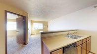 This single-floor space is designed with an open-concept layout that seamlessly connects the kitchen and living area, creating a functional and cohesive environment. The kitchen is equipped with a double stainless steel sink, a dishwasher, and warm-toned laminate countertops that extend along a breakfast bar-style divider, separating the kitchen from the main living space. Cabinetry with a rich wood finish adds a classic touch to the kitchen area. The living area features plush wall-to-wall carpeting in a neutral tone, contributing to a comfortable and inviting atmosphere. A window at the far end of the living space allows natural light to filter in, brightening the room and offering a view of the exterior surroundings. The walls are finished in light, neutral tones, complementing the overall warm palette of the space. A textured ceiling adds subtle visual depth throughout. A doorway visible along one wall provides access to additional areas of the unit. The layout is efficient and well-organized, making practical use of the available space while maintaining an open and airy feel throughout.