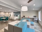 Located at 1100 Indian Trail Lilburn Rd NW in Norcross, Georgia, this thoughtfully designed single-floor space blends modern aesthetics with functional versatility. The interior is unified by striking herringbone-patterned wood-look flooring that flows throughout, complemented by a cohesive teal and neutral color palette that lends a vibrant yet polished atmosphere.

The open social area serves as the heart of the space, featuring a white round table surrounded by teal upholstered chairs with chrome frames, alongside a built-in island with bar seating and modern globe pendant lighting. A bold teal ceramic tile accent wall anchors the kitchenette, which is outfitted with a retro-style mint green SMEG refrigerator, a coffee station, and white cabinetry — adding a playful, curated touch.

The lounge zone offers tiered modular seating in teal and gray, positioned atop a multi-toned area rug and oriented toward a wall-mounted flat-screen TV. Large windows flood this area with natural light and frame views of the surrounding greenery and outdoor amenities. A dedicated workspace area provides desk stations and ergonomic seating, ideal for productivity. The space connects seamlessly to additional rooms, including what appears to be a formal sitting room, creating a well-rounded and inviting environment throughout.
