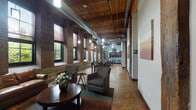 Located on Mississippi Avenue in St. Louis's vibrant Tower Grove South neighborhood, this thoughtfully designed single-floor space masterfully combines industrial heritage with contemporary comfort. The open floor plan is anchored by soaring ceilings with exposed wooden beams and visible ductwork, lending the interior a raw, loft-like aesthetic that feels both expansive and welcoming.

Original exposed brick walls line the perimeter, punctuated by a series of tall, large-pane windows that flood the space with abundant natural light and frame views of the surrounding greenery. Warm, richly patterned hardwood flooring runs throughout, tying the various zones together cohesively.

The lounge area features a leather sofa, upholstered armchairs, a round wooden coffee table, and a wall-mounted flat-screen TV, creating an inviting space for relaxation or casual gatherings. Adjacent to the lounge, a dedicated dining area accommodates a round glass-top table with seating for multiple guests.

The kitchen is outfitted with dark espresso cabinetry, granite countertops, stainless steel appliances, and a breakfast bar with bar-height seating, all illuminated by pendant lighting suspended from the lofty ceiling. Multiple private rooms with dark-framed doors provide additional functional space. The overall ambiance is one of refined industrial elegance, balancing historic architectural character with modern, purposeful design.