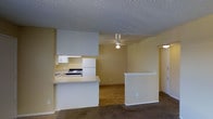 This single-floor unit offers a practical and functional layout across several interconnected spaces. The open-concept design connects the kitchen directly to the living area, creating a cohesive flow between the two spaces. The kitchen is equipped with white cabinetry, a gas range, a full-size refrigerator, and a sink, all complemented by a peninsula counter that serves as a natural divider between the kitchen and living room. A textured ceiling and a ceiling fan with a light fixture add character to this shared space.

The living area features warm-toned carpet flooring, while the kitchen and adjoining spaces transition to hardwood-style flooring, adding visual contrast throughout the unit. A half-wall partition near the entryway provides subtle definition between areas without compromising the open feel of the layout.

The bathroom is outfitted with standard fixtures, including a toilet, sink, and bathtub, and is conveniently positioned off the main corridor. Additional rooms, visible in the floor plan, offer flexible use as bedrooms or private spaces. Natural light filters into the unit, contributing to a bright and welcoming overall ambiance.
