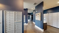 This single-floor mail and parcel center is designed to serve a residential community with both functionality and visual cohesion. The space is immediately distinguished by its bold navy blue walls, which are offset by crisp white baseboards and a light ceiling, creating a striking yet polished aesthetic throughout. Wood-look tile flooring in a warm gray-brown tone runs consistently across the entire area, reinforcing a modern, durable design sensibility.

The room is organized into clearly defined zones. A dedicated wall of individual resident mailboxes, finished in brushed silver, lines one section, offering numbered compartments for organized mail retrieval. Adjacent to this, a large bank of smart package lockers provides secure, automated parcel storage, accommodating the demands of modern delivery services. A rent-and-return tool kiosk adds a distinctive, resident-focused amenity, stocking everyday tools available for borrowing.

Recessed ceiling lighting and wall-mounted sconces ensure the space is evenly and adequately illuminated. Large windows in select areas allow natural light to enter, offering views of surrounding greenery. The layout is open and navigable, with structural columns serving as design elements that break up the space without impeding flow. Overall, the room balances practical utility with a clean, contemporary aesthetic.