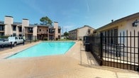 Located in Sherman, this outdoor community pool area serves as a central amenity within a multi-building residential complex. The centerpiece is a large, irregularly shaped swimming pool filled with clear, blue-green water, offering ample space for recreation and leisure. The pool is surrounded by a spacious aggregate concrete sundeck, providing room for lounge chairs and relaxation under the open Texas sky.

The pool area is fully enclosed by sturdy black wrought iron fencing, ensuring security and controlled access through gated entries. Adjacent to the pool, a single-story brick facility building houses essential amenities, including restrooms and utility access, constructed with classic tan brick and a pitched roof. Posted pool hours and rules are clearly displayed on the exterior of this building.

The surrounding complex features two-story residential buildings with stucco and brick exteriors, covered balconies, decorative chimneys, and exterior staircases, all framed by mature trees that provide natural shade and a pleasant backdrop. Well-maintained grassy areas and paved pathways connect the various sections of the property. The overall setting offers a practical and inviting outdoor environment, ideal for warm-weather recreation in a community-centered atmosphere.