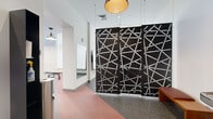 Located at 31 Elm St in Springfield, MA, this fitness center offers a thoughtfully designed, single-floor space that balances functionality with visual appeal. The layout is generously proportioned, providing ample room for a diverse range of workout equipment, including treadmills, ellipticals, a recumbent bike, cable machines, free weights, and agility bars.

A standout feature of the space is a large, vivid wall mural depicting sweeping abstract landscapes in rich tones of orange, blue, and earth, lending an energizing artistic focal point to the workout area. Structural columns finished in deep black punctuate the open floor plan while complementing the industrial-modern aesthetic.

Flooring transitions between speckled rubber and coral-toned runners guide movement throughout the space. Pendant lighting with bell-shaped shades provides warm, even illumination throughout. Large windows along the perimeter draw in natural light and offer views of the surrounding urban streetscape.

An entryway area features striking black decorative screen panels adorned with a geometric dot-and-line pattern, creating a stylish visual boundary. Additional amenities include a mirrored wall section and informational signage. The overall ambiance is modern, motivating, and well-suited for an active fitness environment.