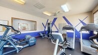 Located at 9501 Short Spoon Ct in Glen Allen, Virginia, this fitness center offers a comprehensive workout environment within a single, efficiently organized room. The space is defined by its eye-catching design: crisp white walls accented by bold cobalt blue geometric stripe patterns, creating a dynamic and energetic atmosphere. Dark rubber flooring runs throughout, providing durability and traction suited to a high-use exercise environment. A textured ceiling with flush-mounted lighting panels and a ceiling fan ensures the space remains well-lit and ventilated during workouts.

Large windows along one wall admit generous natural light while offering views of the surrounding outdoor greenery, enhancing the overall ambiance. A large wall mirror expands the sense of space and allows users to monitor their form. Equipment is thoughtfully arranged to maximize the available floor area and includes Precor cardio machines such as ellipticals, a rowing machine, treadmills, and a stationary bike, alongside a multi-station cable strength machine, free weight dumbbells, and adjustable benches. A bulletin board with workout guides and a wall clock add practical, informational touches. The entry features a classic wood-framed glass door, contributing a warm architectural detail to the otherwise modern, athletic interior.