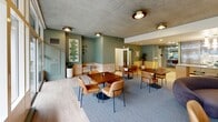Located at 123 NW 12th Ave in Portland's vibrant Pearl District, this thoughtfully designed amenity space combines modern aesthetics with functional versatility. The single-floor layout is organized into three distinct areas that flow seamlessly together, offering residents a welcoming environment for socializing, dining, and relaxing.

The open lounge area is anchored by a large area rug and furnished with a mix of curved sofas and warm-toned leather chairs arranged around low tables, creating an inviting gathering atmosphere. A striking tiled accent wall serves as a dramatic focal point, featuring a linear fireplace and wall-mounted television — perfect for both cozy evenings and casual entertainment.

Floor-to-ceiling glass windows and sliding doors line one wall, flooding the space with abundant natural light and providing direct views of the outdoor courtyard and surrounding greenery. The herringbone-patterned light wood flooring transitions seamlessly throughout the lounge areas, adding warmth and texture.

The gourmet kitchen is a standout feature, outfitted with sleek high-gloss cabinetry in a sophisticated slate blue, accented with brushed gold hardware. White quartz countertops, a marble-look tile backsplash, under-cabinet lighting, a wine cooler, and premium stainless steel appliances elevate the space. A large central island with fluted wood paneling and bar seating serves as both a prep area and a social hub, finished with elegant globe pendant lights above.