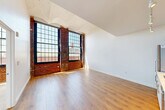 Located at 255 Promenade St in Providence, Rhode Island, this one-bedroom, one-bathroom loft-style unit showcases a compelling blend of historic industrial character and modern finishes. The open-plan living, dining, and kitchen area is the heart of the space, distinguished by soaring ceilings with arched detailing, warm hardwood flooring, and dramatic floor-to-ceiling multi-pane windows framed by exposed red brick columns. These oversized windows flood the interior with abundant natural light and offer sweeping views of the Providence skyline. The kitchen is thoughtfully designed with light wood cabinetry, open shelving, stainless steel appliances including a refrigerator and dishwasher, a sleek backsplash, and track lighting overhead. The spacious primary bedroom continues the industrial aesthetic with exposed brick accents, large industrial-style windows, and generous closet space provided by two dedicated closets. The bathroom is finished with elegant marble-style tile throughout, featuring a glass-enclosed walk-in shower for a clean, contemporary feel. Throughout the unit, the juxtaposition of raw brick and polished modern elements creates a distinctive urban ambiance that is both functional and visually striking.