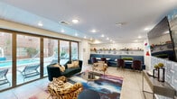 Located at 595 King St in the heart of Charleston, SC, this stylish community lounge is a single-floor gathering space designed for comfort and social connection. The open layout is anchored by a bold, colorful area rug that sets the tone for the eclectic décor throughout. Plush seating — including a deep teal velvet sofa and animal-print accent chairs — is arranged around a glass-top coffee table, creating an inviting lounge area. A wall-mounted flat-screen TV is flanked by a textured geometric tile feature wall, while neon wall lettering adds a fun, contemporary character to the space. A dedicated dining area with a marble-top table and upholstered chairs sits adjacent to the lounge, backed by display shelving lined with decorative plates and greenery. The kitchenette area features a striking marble-topped peninsula with bar seating, pendant lighting, built-in cabinetry, and a glass block accent wall that diffuses soft, ambient light. Warm wood built-in open shelving provides additional display and storage. Large glass French doors open directly to an outdoor pool terrace, flooding the space with natural light and offering seamless indoor-outdoor connectivity. Light tile flooring and recessed ceiling lighting complete the clean, modern aesthetic throughout.
