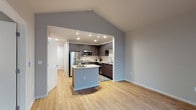 This single-floor residence showcases a thoughtfully designed open-concept layout that seamlessly connects the living area and kitchen. The space is immediately distinguished by its vaulted ceilings, which lend an airy, expansive quality to the main living area. Light hardwood flooring runs throughout the common spaces, contributing to a warm and cohesive aesthetic.

The kitchen is a standout feature, equipped with dark espresso-toned cabinetry that contrasts elegantly against light granite countertops. A central island with a curved granite surface provides additional workspace and casual seating. Stainless steel appliances—including a refrigerator, range, and microwave—complete the modern kitchen ensemble. Track lighting and recessed fixtures ensure the kitchen is well-illuminated.

The bathroom continues the home's design language with matching dark cabinetry, a granite-topped vanity with an undermount sink, chrome hardware, and light hardwood-style flooring. A large mirror enhances the sense of space.

The bedroom offers a quieter retreat, finished with plush carpeting, neutral gray walls, and a sliding-door closet that maximizes storage. The overall palette of soft grays and warm wood tones creates a clean, contemporary ambiance throughout the unit.