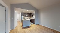 This single-floor residence showcases a thoughtfully designed open-concept layout that seamlessly connects the living area and kitchen. The space is immediately distinguished by its vaulted ceilings, which lend an airy, expansive quality to the main living area. Light hardwood flooring runs throughout the common spaces, contributing to a warm and cohesive aesthetic.

The kitchen is a standout feature, equipped with dark espresso-toned cabinetry that contrasts elegantly against light granite countertops. A central island with a curved granite surface provides additional workspace and casual seating. Stainless steel appliances—including a refrigerator, range, and microwave—complete the modern kitchen ensemble. Track lighting and recessed fixtures ensure the kitchen is well-illuminated.

The bathroom continues the home's design language with matching dark cabinetry, a granite-topped vanity with an undermount sink, chrome hardware, and light hardwood-style flooring. A large mirror enhances the sense of space.

The bedroom offers a quieter retreat, finished with plush carpeting, neutral gray walls, and a sliding-door closet that maximizes storage. The overall palette of soft grays and warm wood tones creates a clean, contemporary ambiance throughout the unit.