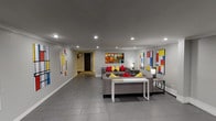 This thoughtfully designed single-floor space serves as a multifunctional common area, blending comfort, art, and utility in a cohesive layout. The entrance greets visitors with a welcoming foyer, complete with glass doors, a decorative console table, a geometric sculpture, and a potted plant — establishing an inviting first impression.

The lounge area is the centerpiece of the space, featuring multiple gray upholstered sofas accented with bold red and yellow throw pillows. A glass-top coffee table with a geometric wood base anchors the seating arrangement, while wall-mounted lamps provide warm, ambient lighting. The gray tile flooring runs throughout, complementing the neutral gray walls and white crown molding that add a clean, polished finish.

A striking collection of large-scale Mondrian-inspired artworks — bold geometric compositions in primary colors — lines the walls, lending the space an energetic, gallery-like character. Additional abstract artwork adds further visual interest.

Toward the rear of the space, fitness equipment is visible, indicating a dedicated workout zone. Recessed ceiling lighting throughout ensures the entire floor is well-illuminated. The overall ambiance is modern and curated, balancing artistic expression with practical, comfortable amenities suited for a communal setting.