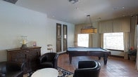 This single-floor space is designed as a stylish recreational lounge, offering a well-appointed environment for entertainment and relaxation. The centerpiece of the room is a full-size pool table finished in dark wood with a slate-blue felt surface, illuminated by a statement rectangular pendant light with a warm-toned shade that adds a classic, sophisticated touch overhead. The flooring consists of terracotta tiles that lend a warm, earthy character to the space, partially accented by a decorative area rug beneath the seating arrangement. Comfortable black leather club chairs are arranged around a round side table, creating an intimate lounge area ideal for socializing. Natural light filters into the room through windows dressed with neutral drape panels and blinds, contributing to a bright and welcoming ambiance. The walls are finished in a clean white, complemented by subtle recessed lighting throughout the ceiling. A pair of tall, rustic wooden decorative panels mounted on one wall add an architectural focal point with a touch of old-world charm. A vintage-style wooden sideboard with decorative items completes the room's character. The overall aesthetic balances casual comfort with refined design details, making it a versatile and engaging communal space.