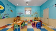 This single-level children's playroom is designed with a captivating underwater theme that envelops the entire space in a sense of whimsy and discovery. The walls are painted in a bright aqua blue and adorned with detailed hand-painted murals depicting an array of marine life, including dolphins, orca whales, tropical fish, an octopus, a sea turtle, and a cheerful yellow submarine. A painted seagrass border runs along the lower portion of the walls, creating a cohesive oceanic narrative throughout the room.

The floor features a bold, multicolored checkerboard pattern in shades of blue, yellow, red, and white, adding a playful energy underfoot. Child-sized rectangular and round tables are paired with brightly colored chairs in green, red, blue, and yellow, providing ample space for group activities and creative play. Open wooden shelving units along the walls offer organized storage for toys and educational materials.

The room benefits from natural light streaming through large windows and glass French doors, which also provide outdoor views and easy access to exterior areas. Recessed lighting and a ceiling fan ensure comfortable illumination and airflow. The overall ambiance is energetic, imaginative, and purposefully designed to engage and delight young children.