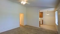 This single-floor residence offers a straightforward, functional layout designed for comfortable everyday living. The main living area is finished with wall-to-wall carpet and painted in soft, neutral tones that lend a calm and airy atmosphere throughout. A ceiling fan with a built-in light fixture is centrally mounted overhead, providing both ventilation and ambient lighting. The living space connects seamlessly to the kitchen, which is partially open to the main area via a pass-through wall. The kitchen features warm-toned wooden cabinetry, light countertops, and a dishwasher, offering practical storage and workspace. A hallway accessible from the living area leads to additional rooms within the residence, suggesting a well-organized floor plan. The clean, understated aesthetic of the space makes it well-suited for a variety of furnishing styles. Neutral wall colors, consistent flooring materials, and a simple yet cohesive design language give the home a move-in-ready feel. The layout prioritizes ease of movement and accessibility between common areas, making the space practical for daily routines.