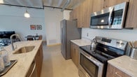 This one-bedroom, one-bathroom residence at Checker showcases an industrial-chic aesthetic rooted in the building's converted character. The open-concept living and kitchen area is anchored by exposed ceiling infrastructure — including visible ductwork, piping, and track lighting — lending the space a raw, loft-like energy. Large steel-framed factory windows flood the living room with natural light while framing urban views, and original brick accent walls add warmth and texture to the interior.

The kitchen is thoughtfully appointed with warm wood cabinetry, light stone countertops, and a full suite of stainless steel appliances including a refrigerator, electric range, and over-the-range microwave. A generous island with seating provides both prep space and a casual dining area, seamlessly connecting the kitchen to the open living zone. Polished concrete-style flooring runs throughout, tying the spaces together cohesively.

The bedroom offers a calm retreat with a ceiling fan, closet storage, and an exposed ceiling that maintains the industrial character found throughout the unit. The full bathroom features a wide vanity with stone countertop, rich wood cabinetry, a large mirror, and clean, neutral finishes. This unit delivers a distinctive urban living experience with strong design continuity from room to room.