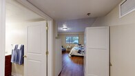 This single-floor residential unit offers a well-organized and comfortable living environment designed with both functionality and accessibility in mind. The bedroom is generously proportioned, accommodating multiple beds and seating, and features warm hardwood-style flooring, a textured ceiling, and a window that allows natural light to fill the room while providing views of the surrounding exterior. A white wardrobe unit offers ample storage, and decorative wall art adds a welcoming touch to the neutral, cream-toned walls. A private entryway with white panel doors leads into the bedroom area, enhancing the sense of separation and privacy. The bathroom is thoughtfully designed with accessibility as a priority, featuring grab bars, a roll-in shower with a curtained enclosure, a toilet, and a wood-vanity with an integrated sink and mirror. The hardwood-style flooring continues seamlessly into this space. The compact kitchen is efficiently laid out with white cabinetry extending to the ceiling, a gas range, a full-size refrigerator, and a stainless sink set into a gray countertop. Under-cabinet lighting provides practical illumination. Throughout the unit, the consistent warm flooring, neutral palette, and clean finishes create a cohesive, calm, and livable atmosphere.