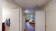 This single-floor residential unit offers a well-organized and comfortable living environment designed with both functionality and accessibility in mind. The bedroom is generously proportioned, accommodating multiple beds and seating, and features warm hardwood-style flooring, a textured ceiling, and a window that allows natural light to fill the room while providing views of the surrounding exterior. A white wardrobe unit offers ample storage, and decorative wall art adds a welcoming touch to the neutral, cream-toned walls. A private entryway with white panel doors leads into the bedroom area, enhancing the sense of separation and privacy. The bathroom is thoughtfully designed with accessibility as a priority, featuring grab bars, a roll-in shower with a curtained enclosure, a toilet, and a wood-vanity with an integrated sink and mirror. The hardwood-style flooring continues seamlessly into this space. The compact kitchen is efficiently laid out with white cabinetry extending to the ceiling, a gas range, a full-size refrigerator, and a stainless sink set into a gray countertop. Under-cabinet lighting provides practical illumination. Throughout the unit, the consistent warm flooring, neutral palette, and clean finishes create a cohesive, calm, and livable atmosphere.