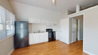 This single-floor space is defined by its distinctive vaulted ceilings, which lend an airy, open feel to the interior. Warm hardwood flooring runs consistently throughout, tying each area together with a clean, cohesive look.

The kitchen is well-equipped with ample white cabinetry both above and below the counter, offering generous storage space. It includes a stainless single-basin sink, a black electric range with a ventilation hood above, and a full-size refrigerator. The light countertops and white finishes give the kitchen a bright, functional character.

Adjacent to the kitchen is an open living area bathed in natural light, courtesy of multiple windows fitted with horizontal blinds. A glass-panel exterior door leads to the outside, further enhancing the sense of openness and connecting the interior to the outdoors. A wall-mounted HVAC unit provides climate control for the space.

The bedroom area benefits from the same soaring vaulted ceiling, creating a spacious and comfortable environment. Neutral-toned walls throughout contribute to the calm, understated ambiance of the space.

The bathroom is straightforwardly appointed with a full-size soaking tub, a toilet, and textured walls, providing all essential amenities in a compact, efficient layout. Hardwood flooring extends into the bathroom as well, maintaining the design continuity seen across the rest of the space.