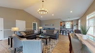 Located at 35 Hidden Oak Way in Manchester, New Hampshire, this single-floor space offers a thoughtfully designed open-concept layout that seamlessly connects the living, dining, and kitchen areas. The main living area is anchored by soaring vaulted ceilings and a striking wagon-wheel chandelier, creating a bright and airy atmosphere. Warm-toned hardwood-style flooring flows throughout, complemented by neutral gray walls and white wainscoting that lend a clean, cohesive aesthetic. Recessed lighting and large windows invite generous amounts of natural light, with views of lush greenery outside. The living area provides ample space for comfortable seating arrangements, with a classic fireplace visible as a central focal point. Adjacent to the living area, a dedicated dining space features large multi-pane windows that frame picturesque outdoor views, making it an ideal setting for gatherings. The kitchen is equipped with rich medium-toned wood cabinetry, granite countertops, a peninsula with bar seating, and black appliances, with a pendant light adding a refined touch. The overall ambiance of the space is welcoming and polished, balancing modern finishes with a warm, inviting character throughout.