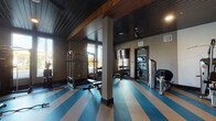 This fitness center is thoughtfully designed to accommodate a wide range of workout styles within a single-floor layout. The space is divided into distinct functional zones, each tailored to specific training needs. The main strength training area features a vaulted, dark wood-paneled ceiling supported by exposed beams, lending the room a refined yet energetic atmosphere. Bold blue and tan striped rubber flooring runs throughout this zone, adding a dynamic visual element while providing durable, impact-absorbing underfoot support. Large windows along multiple walls invite generous natural light and offer views of the surrounding outdoor landscape, enhancing the overall workout environment.

The strength area is well-stocked with a power rack, adjustable weight bench, a full dumbbell rack ranging across multiple weight increments, and cable machines. Full-length mirrors are strategically placed to support form monitoring during training. A dedicated cycling studio adjoins the main floor, featuring multiple stationary bikes, floor-to-ceiling windows, a large wall-mounted television, and a ceiling fan for added ventilation. An additional section houses selectorized resistance machines alongside mirrored walls. Ceiling fans and recessed lighting provide consistent comfort and illumination throughout the entire facility.