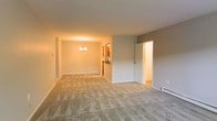 This single-floor residential space offers a straightforward and functional layout designed for comfortable everyday living. The open living and dining area forms the heart of the home, featuring wall-to-wall plush carpeting in a neutral beige tone that adds warmth underfoot and contributes to a cohesive aesthetic throughout. The walls are finished in soft, muted gray-beige tones, creating a calm and versatile backdrop that suits a variety of décor styles. A classic chandelier anchors the dining area, providing both ambient lighting and a subtle decorative focal point. The space transitions naturally between living and dining zones, making it well-suited for both relaxing and entertaining. A baseboard heater along the far wall ensures consistent warmth, reflecting practical residential construction. Doorways lead to additional rooms, including what appears to be a kitchen visible in the background, hinting at a well-connected floor plan. A secondary door opens to another room, likely a bedroom or utility space, reinforcing the home's efficient use of space. Overall, the residence presents a clean, move-in-ready environment with a neutral palette and modest finishes that provide a solid foundation for personalization.
