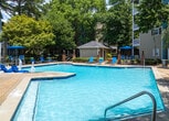 Located at 1570 Sheridan Rd NE in Atlanta, Georgia, this outdoor amenity area centers around a generously sized resort-style swimming pool that serves as the focal point of the space. The pool features an integrated shallow wading area with built-in water fountains and distinctive blue lounger chairs positioned directly in the water, offering a unique and relaxing experience. A dedicated pool deck surrounds the water, furnished with aqua-blue chaise lounges and umbrella-shaded table seating, providing ample space for sunbathing and socializing. A charming pool house structure sits adjacent to the deck, offering additional convenience for residents. The entire area is enclosed by a classic wrought-iron fence accented with brick columns, lending a sense of security and elegance. Mature trees, including a striking weeping willow and a variety of deciduous trees displaying seasonal color, frame the perimeter and provide natural shade throughout the space. The multi-story residential buildings surrounding the pool feature balconies that overlook the amenity area, creating a cohesive and community-oriented outdoor environment. The overall ambiance is bright, inviting, and well-maintained, making it an ideal retreat for relaxation within the heart of Atlanta.