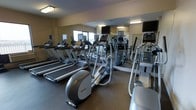 This fitness center offers a focused and functional environment for cardiovascular training and general exercise. The room is lined with a full wall of mirrors, which visually expands the space and allows users to monitor their form during workouts. Overhead fluorescent lighting ensures the area is well-lit throughout.

The cardio equipment lineup includes multiple treadmills arranged along one side of the room, providing ample capacity for users during peak hours. Elliptical cross-trainers are positioned centrally, offering low-impact workout options. Additional cardio machines, including stair climbers and stationary bikes, are visible toward the far end of the room. Each machine is equipped with a personal monitor or display screen, and several wall-mounted flat-screen televisions are positioned throughout the space for entertainment during workouts.

The flooring features a durable, dark rubberized surface suited for high-traffic fitness use. A window along one wall introduces natural light into the space. The overall layout is practical and efficient, with equipment arranged to maximize usability while maintaining comfortable circulation space between machines.