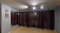 This yoga room offers a calm, purposefully designed studio space ideal for practice and instruction. The open floor plan is finished with smooth, light-toned hardwood-style flooring that provides a clean and supportive surface for yoga sessions. Soft blue-gray walls create a tranquil atmosphere throughout the room, contributing to a peaceful and focused environment.

A standout feature is the extensive use of deep burgundy sheer curtains, which line much of the perimeter and partially conceal large wall mirrors behind them. These mirrors allow practitioners to observe and correct their form, while the curtains add a layer of privacy and an intimate ambiance. Track lighting installed on the textured ceiling provides adjustable, directed illumination across the studio.

The entry features a set of warm wood-toned double doors, flanked by wall-mounted shelving that holds product supplies and accessories. Informational charts and reference materials are displayed on the walls, adding a functional, educational element to the space.

A dedicated corner stores a neatly stacked collection of yoga mats and blankets alongside a wicker storage basket, keeping essentials organized and accessible. Decorative wall accents, including a delicate branch-style wall piece, add a subtle artistic touch. Overall, the room is thoughtfully arranged to support a serene and focused yoga practice.