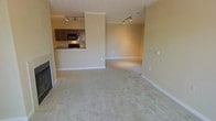 This single-floor residence offers an open and airy layout that maximizes its living space. The main living area is anchored by a tiled fireplace, providing a warm focal point for the room. Plush, neutral-toned carpeting extends throughout the living and adjoining areas, lending a comfortable and cohesive feel to the space. Track lighting mounted on the ceiling provides targeted illumination throughout.

The kitchen is positioned at a slightly elevated counter level, creating a natural separation from the living area while maintaining an open connection to it. It features dark wood cabinetry, a raised breakfast bar that doubles as a casual dining space, and black appliances including a range and an overhead microwave. The countertops offer ample workspace, and tile flooring in the kitchen area provides a practical contrast to the carpeted living zone.

Crown molding lines the ceilings throughout, adding a refined architectural detail that elevates the overall aesthetic. The warm, neutral wall tones throughout the space contribute to a welcoming and versatile ambiance, making it well-suited for a variety of interior styles. The layout flows smoothly between the kitchen and living areas, making it ideal for both everyday living and entertaining.