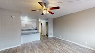 This single-floor unit offers a practical and efficiently organized layout, encompassing a living room, kitchen, bathroom, and hallway within a compact footprint. Light wood-style vinyl flooring runs consistently throughout the space, lending a clean, cohesive look that connects each area visually.

The living room serves as the central space of the unit, featuring neutral gray walls, a textured popcorn ceiling, and a ceiling fan with an integrated light fixture. A wall-mounted air conditioning unit and a window with blinds provide climate control and natural light. A closet accessible from the living area offers convenient built-in storage.

The kitchen is positioned adjacent to the living room in an open-plan arrangement, separated by a peninsula counter finished with a gray speckled laminate surface. White cabinetry provides ample storage above and below, complemented by a tiled backsplash, a double-basin sink, and a black electric range. A textured ceiling and flush-mount lighting complete the kitchen's functional design.

The bathroom features wood-style tile flooring, a vanity with a speckled countertop and under-sink cabinetry, a wall-mounted mirror with lighting, and a separate room housing the toilet and a full bathtub with a shower surround. The hallway connects the main living areas to the bathroom and includes a closet, maintaining an organized flow throughout the unit.
