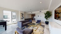 Located on Melrose Avenue in Los Angeles, this single-floor lobby and fitness center combines contemporary design with upscale finishes to create a welcoming and functional communal space. The lobby immediately impresses with its polished marble-look tile flooring, bold navy and gold abstract wallcoverings, and plush deep-blue velvet seating including a sweeping curved sofa and barrel chairs with gold-toned bases. A round marble-top coffee table anchors the lounge area, while floor-to-ceiling glass windows flood the space with natural light and offer open views of the tree-lined streetscape outside. A built-in workstation with a bar-height counter and rounded seating provides a practical workspace alongside the lounge. The elevator foyer features clean white paneled walls, a contrasting dark textured accent wall, and a sculptural circular chandelier that adds a refined focal point overhead. The on-site fitness center is thoughtfully equipped with TRUE brand cardio machines including treadmills, ellipticals, and stationary bikes, as well as a full dumbbell rack, a cable machine, an adjustable bench, and a stability ball. Motivational artwork and a wall-mounted television complete the gym environment. Throughout, recessed lighting and consistent marble-look flooring tie the spaces together cohesively.