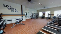 This fitness center is designed to inspire and accommodate a wide range of workout routines within a bright, open layout. The space features durable wood-tone flooring that spans the expansive workout area, providing a clean and energetic environment for exercise. Overhead, ceiling fans ensure consistent airflow and comfort throughout the room, complemented by recessed lighting that keeps the space well-lit at all times. Natural light filters in through windows along the perimeter, adding to the open and airy atmosphere. A bold graphic mural in navy blue, gold, and white reading "Work. Play." adorns one wall, setting a motivating tone for users. The facility is outfitted with a variety of strength training equipment, including a multi-station cable machine and benches, offering versatile options for both resistance and functional training. Built-in white shelving along one wall provides convenient storage for personal items or accessories. French doors connect to an adjacent room, suggesting additional amenities or lounge space nearby. A mounted flat-screen television adds an entertainment element, making workouts more engaging. The overall design is clean, purposeful, and well-organized, creating a welcoming fitness environment.